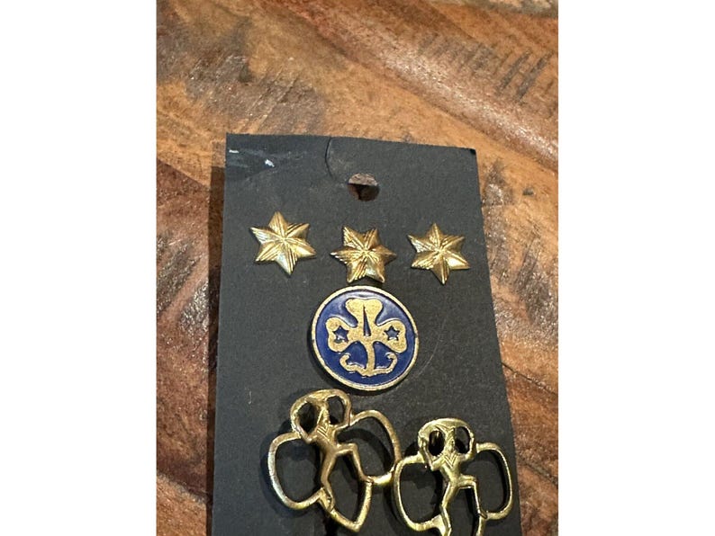 May include: A black card displays Girl Scout pins and badges. Three gold star pins are at the top. Below are a blue and gold round badge and two gold Girl Scout silhouette pins. The items are on a wooden surface.