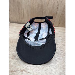 May include: A five-panel cap shown from an interior perspective, featuring a solid black brim and a black strap closure. The interior crown displays a colorful geometric pattern in shades of orange, blue, and white, with a small white brand tag.