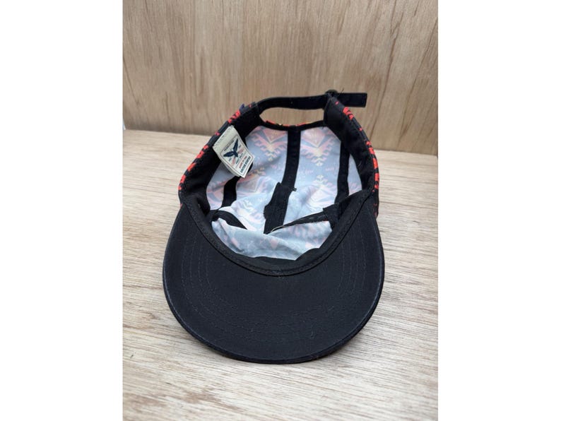 May include: A five-panel cap shown from an interior perspective, featuring a solid black brim and a black strap closure. The interior crown displays a colorful geometric pattern in shades of orange, blue, and white, with a small white brand tag.
