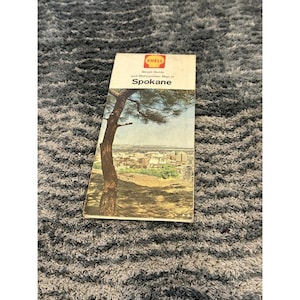 May include: A vintage Shell street guide and metropolitan map of Spokane. The cover features a color photograph of a tree in the foreground overlooking a city skyline. The Shell logo is printed in red and yellow at the top of the white cover.