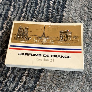 Puede incluir: Una caja rectangular blanca con la etiqueta Parfums de France Sélection 21. La parte superior presenta un panel dorado con ilustraciones en tinta negra de Notre Dame, la Torre Eiffel y el Arco del Triunfo, acentuado por una franja roja, blanca y azul.