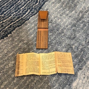 May include: A wooden cribbage board with a leather top and a booklet titled "The Game of Cribbage". The board is brown with small holes for pegs. The booklet is open, displaying text.