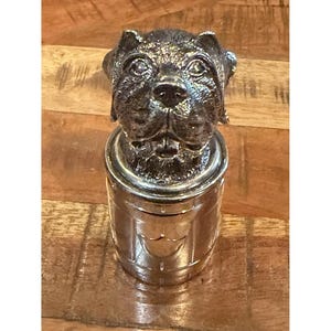 Könnte beinhalten: Ein silberfarbener Flaschenverschluss aus Metall mit einer detaillierten, strukturierten Skulptur eines Hundekopfes an der Oberseite. Der Sockel des Verschlusses ist zylindrisch mit vertikalen Paneelen und einem kleinen schildförmigen Emblem.