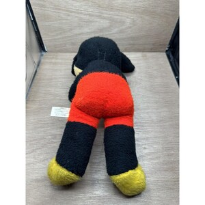 May include: A vintage Mickey Mouse plush toy, lying on its stomach. The toy features a black head and legs, a red torso, and yellow feet. The plush is worn, with visible signs of age.