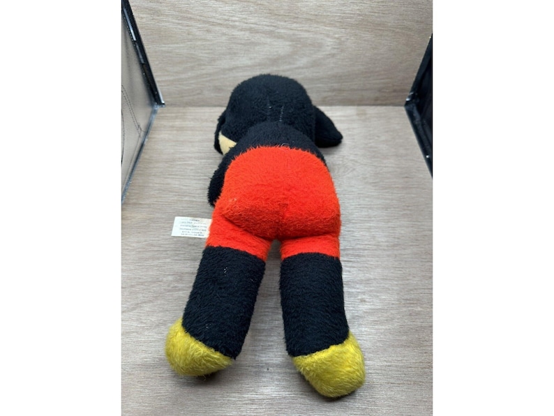 May include: A vintage Mickey Mouse plush toy, lying on its stomach. The toy features a black head and legs, a red torso, and yellow feet. The plush is worn, with visible signs of age.