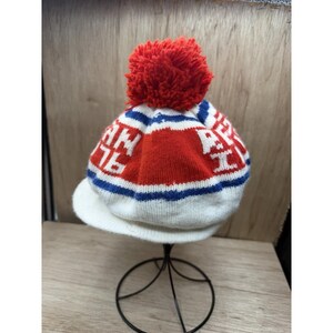 K&ouml;nnte beinhalten: Eine gestrickte Winterm&uuml;tze mit einem roten Bommel. Die M&uuml;tze hat einen wei&szlig;en Schirm und ein Design mit roten, wei&szlig;en und blauen Streifen sowie dem Text "AN 76".