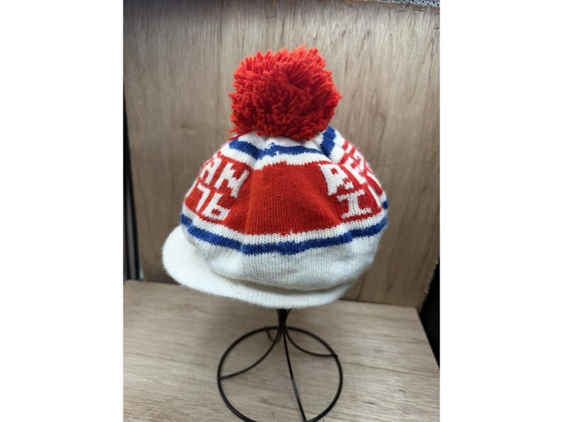 K&ouml;nnte beinhalten: Eine gestrickte Winterm&uuml;tze mit einem roten Bommel. Die M&uuml;tze hat einen wei&szlig;en Schirm und ein Design mit roten, wei&szlig;en und blauen Streifen sowie dem Text "AN 76".