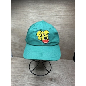 Puede incluir: Gorra de béisbol turquesa con un rostro de perro de dibujos animados amarillo bordado en la parte delantera. El perro tiene la lengua roja fuera. La gorra se exhibe en un soporte de metal negro.