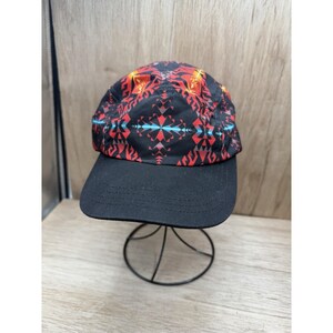May include: A five-panel baseball cap featuring a black base with a vibrant geometric pattern in red, orange, and light blue. The cap has a solid black brim with visible stitching and a structured crown, displayed on a thin black wire stand.