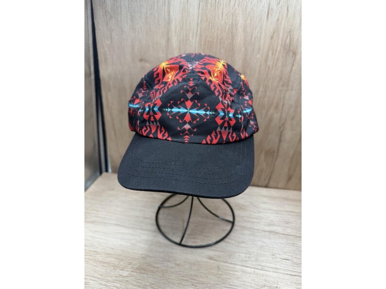 May include: A five-panel baseball cap featuring a black base with a vibrant geometric pattern in red, orange, and light blue. The cap has a solid black brim with visible stitching and a structured crown, displayed on a thin black wire stand.