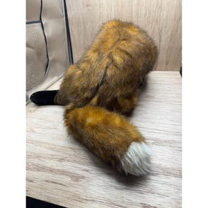 May include: A soft toy fox, featuring a brown and orange body and a white-tipped tail. The toy has a black tail base and is placed on a light-coloured wooden surface.