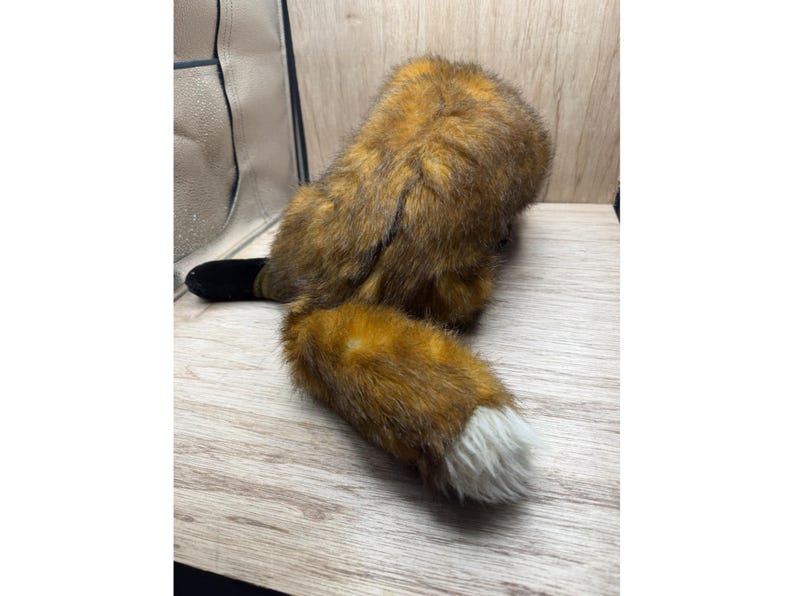 May include: A soft toy fox, featuring a brown and orange body and a white-tipped tail. The toy has a black tail base and is placed on a light-coloured wooden surface.