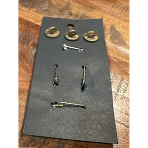 May include: A set of three gold-toned hat pins, a gold-toned lapel pin, and a pair of gold-toned hoop earrings, all mounted on a black card. The pins and earrings are displayed against a wooden surface.