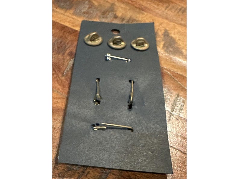 May include: A set of three gold-toned hat pins, a gold-toned lapel pin, and a pair of gold-toned hoop earrings, all mounted on a black card. The pins and earrings are displayed against a wooden surface.