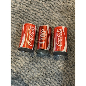 May include: Three Coca-Cola cans. The cans are red with white script and a white curved stripe. The cans are 355 ml.