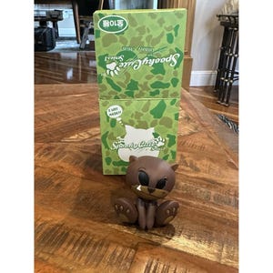 May include: A brown Spooky Cute Series 1 bear figurine with a grumpy expression, sitting in front of two green camouflage boxes. The boxes have the words "Spooky Cute" and "I love music!" printed on them.