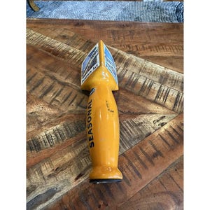 May include: A yellow beer tap handle with the word "SEASONAL" in blue lettering. The top of the tap handle has a label with the words "HARVEST MOON PUMPKIN ALE". The handle is on a wooden surface.