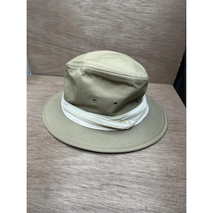 Può includere: Un cappello color cachi con tesa larga e fascia bianca. Il cappello ha una corona arrotondata con fori di ventilazione e un dettaglio con cerniera bianca attorno alla base della corona. Il cappello è su una superficie di legno.