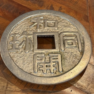 以下が含まれることがあります： 中央に正方形の穴と漢字が刻まれた、丸い銀色の金属製のコイン。 コインには隆起した縁があり、木製の表面に置かれています。 文字は伝統的なフォントです。