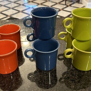 Op de afbeelding: Een verzameling van zeven keramische mokken in drie verschillende kleuren: twee feloranje, twee diepblauw en drie limoengroen. Elke mok heeft een gladde, glanzende afwerking, een cilindrisch lichaam en een klein, rond handvat aan de zijkant.
