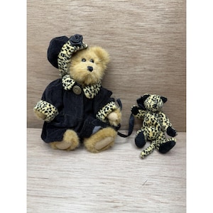 May include: A tan plush teddy bear wearing a black coat with leopard print trim and a matching hat, accompanied by a smaller plush leopard print animal toy with black paws and a long tail, both featuring soft fur and stitched facial details.
