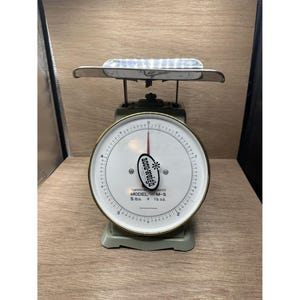 Op de afbeelding: Vintage Acme-Weight weegschaal, model M-5, met een witte wijzerplaat en zwarte markeringen. De weegschaal heeft een zilveren plateau en een groene basis. De schaalverdeling gaat tot 2,27 kg en 340 g.