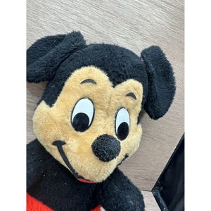 May include: A plush Mickey Mouse toy with a black head, ears, and arms, a tan face, and a red and black body. The toy has large white eyes, a black nose, and a smiling mouth. The toy is lying on a wooden surface.