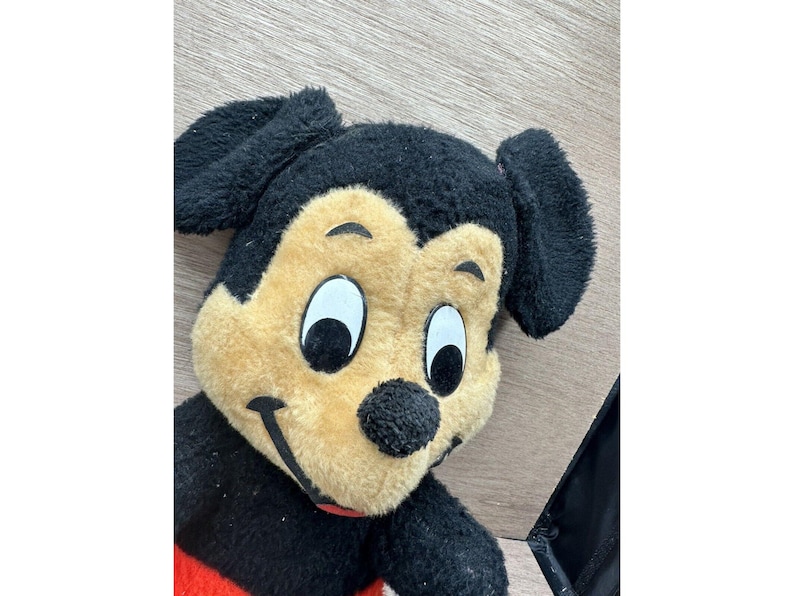 May include: A plush Mickey Mouse toy with a black head, ears, and arms, a tan face, and a red and black body. The toy has large white eyes, a black nose, and a smiling mouth. The toy is lying on a wooden surface.