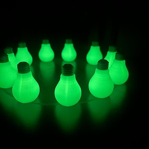 Może przedstawiać: Kolekcja świecących na zielono żarówek ułożonych w okrąg na czarnym tle. Każda żarówka ma ciemnoszarą podstawę i emituje jasne, równomierne zielone światło. Żarówki są równomiernie rozmieszczone, tworząc atrakcyjny wizualnie efekt.