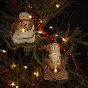 May include: A Christmas tree decorated with a snowman and Santa Claus ornaments. The ornaments are made of fabric and have a rustic, primitive style. The tree is also decorated with red berry garland and white lights.