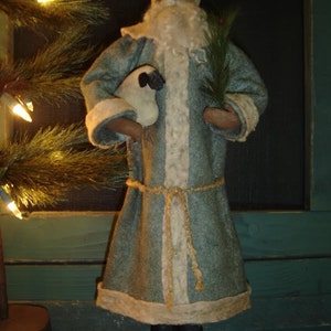 May include: A handmade rag doll dressed as Santa Claus, wearing a blue and white robe with a white fur trim. He is holding a small white lamb in his left arm and a small pine branch in his right hand.