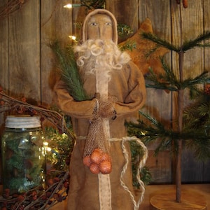 May include: A brown, primitive-style Santa Claus doll with a white beard and a sack of oranges. The doll is standing in front of a wooden background with a small Christmas tree.