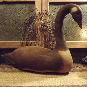 Op de afbeelding: Een bruine en witte primitieve ganzenlokvogel met een lange nek en een klein hoofd. De gans zit op een bruin en beige kleed voor een houten muur met een tinnen paneel.