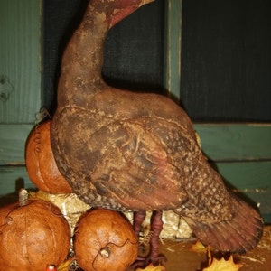 Op de afbeelding: Een bruine en rode primitieve kalkoenfiguur met een blauw oog, zittend op een bed van stro met drie bruine pompoenen.