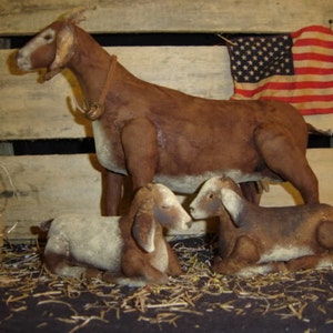 May include: Three brown and white felt goats, a mother and two kids, are posed on a bed of straw. The mother goat is standing with her head turned to the side, while the two kids are lying down. The goats are all made of felt and have a rustic, primitive look.