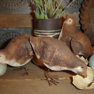 Puede incluir: Tres pájaros de estilo primitivo hechos de tela y alambre, de color marrón y blanco, sentados sobre una superficie de madera con una cáscara de huevo rota y huevos azules.