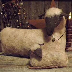 Puede incluir: Figura rústica de oveja con un cordero, hecha de un material beige claro con caras y orejas de color marrón oscuro. Una pequeña campana cuelga del cuello de la oveja. La escena está ambientada sobre un fondo de madera con una estrella y una vela.