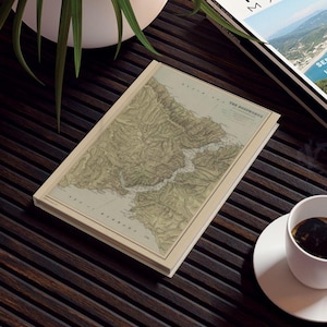May include: A notebook with a vintage map design, in beige, brown, and green tones. The map depicts a coastal region, with the title "THE BOSPHORUS" visible. The notebook is open on a dark wooden surface, next to a cup of coffee.