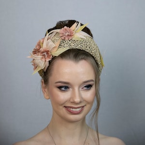 May include: A light brown woven headband with a large pink flower and smaller pink flowers on the side. The headband is worn by a person with light brown hair and makeup.