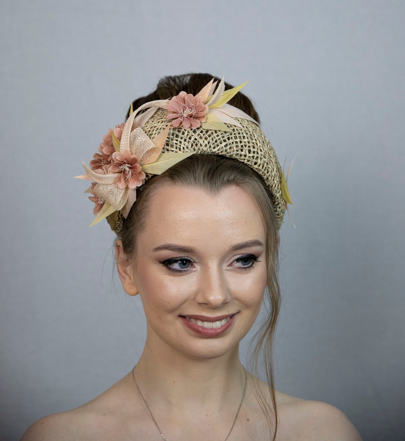 May include: A light brown woven headband with a large pink flower and smaller pink flowers on the side. The headband is worn by a person with light brown hair and makeup.