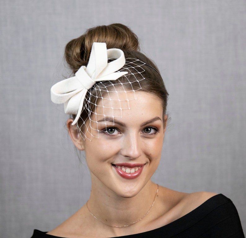 Hellelfenbein oder cremeweißer kleiner Fascinator mit Schleife, wahlweise mit Gesichtsschleier oder auch ohne. Bild 1