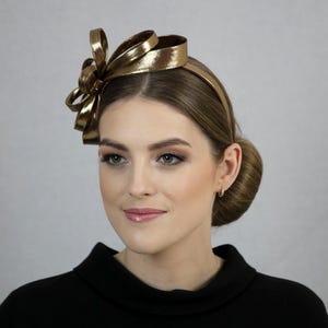 以下が含まれることがあります： 大きなリボンが付いたゴールドカラーのヘアバンド。リボンは光沢のあるメタリック素材で作られています。ヘアバンドは黒いトップスを着た人が着用しています。