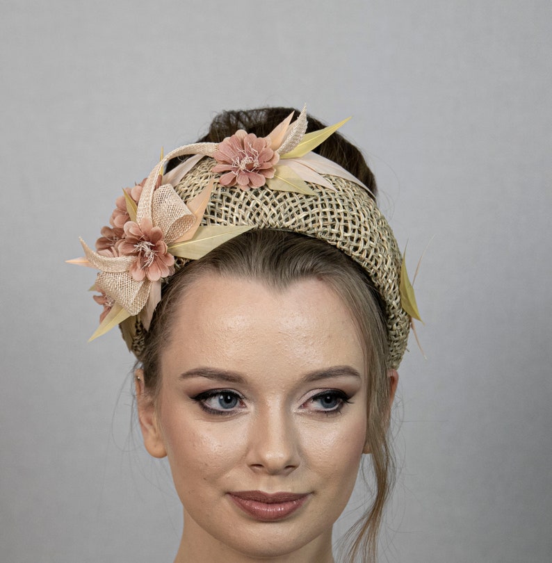 May include: A wide headband made of woven straw with a light brown and beige color scheme. The headband is decorated with pink and beige fabric flowers and leaves.