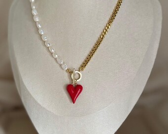 May include: A necklace with a split design. One side features white pearls, the other a gold chain. A red heart pendant hangs from the center. The necklace is displayed on a mannequin.