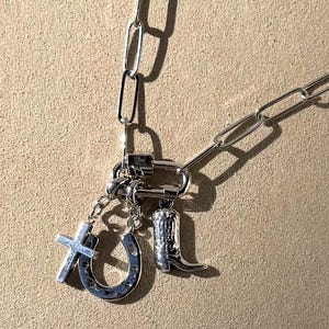 Puede incluir: Un collar de cadena de plata con un cierre de plata y tres dijes: una herradura, una cruz y una bota de vaquero.