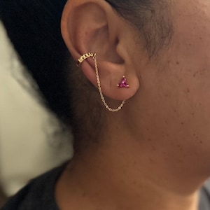 May include: Gold-toned ear cuff with a chain connected to a triangular stud earring with three pink gemstones. The ear cuff wraps around the upper ear, and the stud earring is on the earlobe.