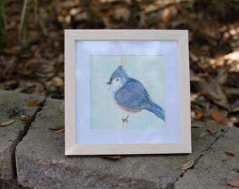 May include: A framed watercolour painting of a bluebird perched on a branch. The bird is facing to the left and has a light blue body with a darker blue head and wings. The background is a light blue.
