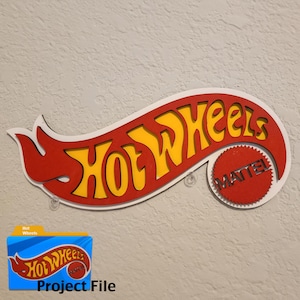 Könnte beinhalten: Ein Holzschild mit dem Hot Wheels-Logo in Rot und Gelb, mit dem Wort "MATTEL" auf einem roten Kreis. Das Schild ist an einer hellgrauen Wand befestigt. Ein blauer Ordner mit dem Hot Wheels-Logo und den Worten "Project File" befindet sich unten links.