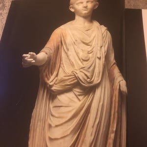 May include: A white marble statue of a young person wearing a toga. The statue is standing with one arm outstretched and the other arm bent at the elbow. The statue is set against a black background.