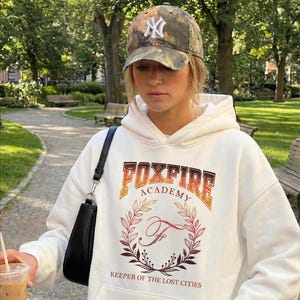 May include: A white hoodie with the text "FOXFIRE ACADEMY" in orange and brown, and a floral design. The person is wearing a camouflage baseball cap with a white logo and carrying a black purse. The background is a park.